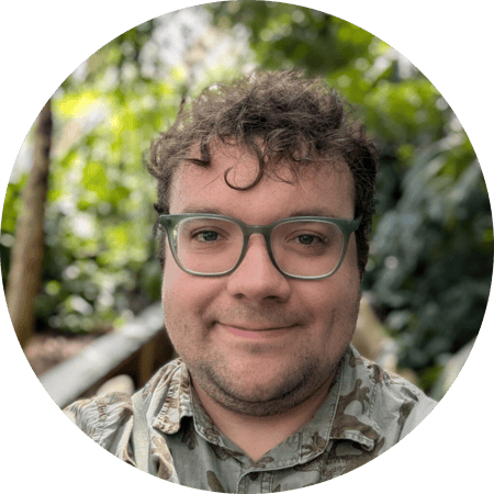 Me wearing a green shirt covered in iguanas and green glasses. I am white, I have brown curly hair, and light facial hair. Blurred greenery is visible in the background.